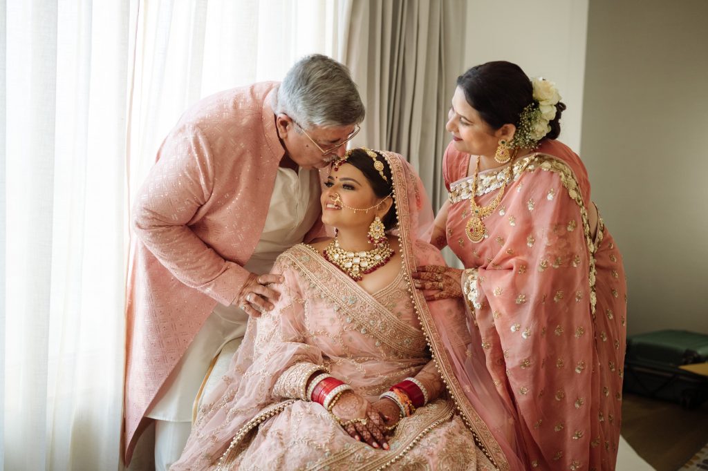 Bride's father kissing on her forehead and mother standing next tot her, all in light pink dress in front of window.